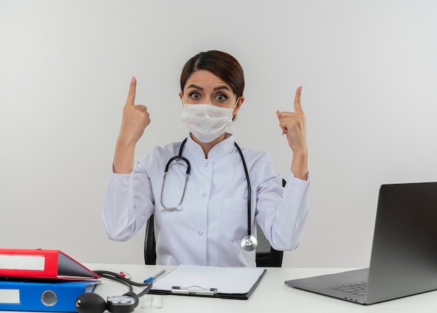 Foto gratuita impressionato giovane dottoressa che indossa abito medico e stetoscopio e maschera medica seduto alla scrivania con strumenti medici e laptop rivolto verso l'alto isolato sul muro bianco