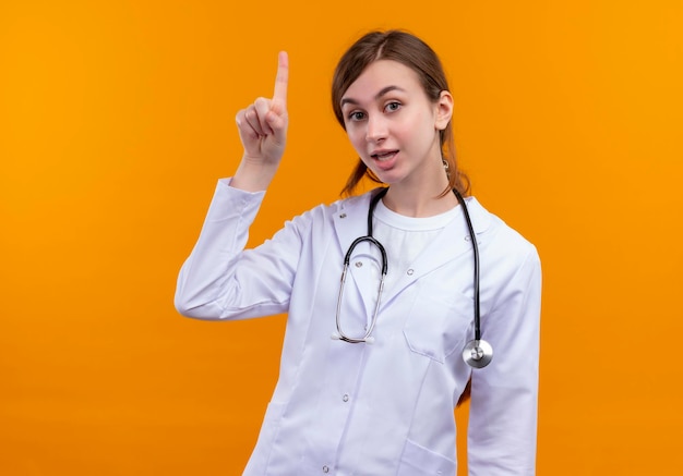コピースペースと孤立したオレンジ色のスペースに指を上げる医療ローブと聴診器を身に着けている感動若い女性医師