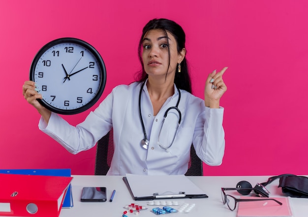 의료용 가운과 청진기를 착용하고 의료 도구가 시계를 들고 분홍색 벽에 고립 된 손가락을 올리는 감동적인 젊은 여성 의사