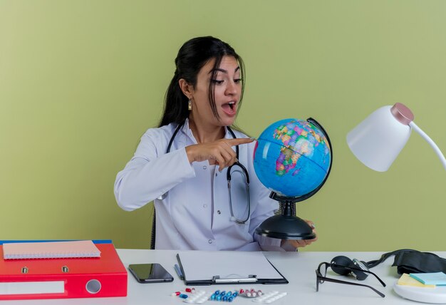 医療用ローブと聴診器を身に着けている感銘を受けた若い女性医師が机に座って医療ツールを保持し、地球を見て、指している