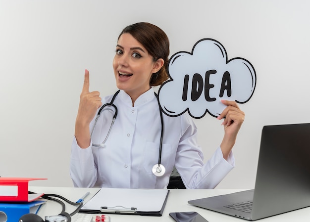 의료 가운과 청진기를 착용하고 의료 도구와 노트북이 흰 벽에 고립 된 아이디어 거품 제기 손가락을 들고 책상에 앉아 감동 된 젊은 여성 의사