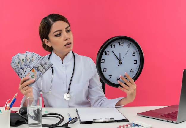 Впечатленная молодая женщина-врач в медицинском халате и стетоскопе сидит за столом с медицинскими инструментами и ноутбуком, держит деньги и часы, глядя на часы