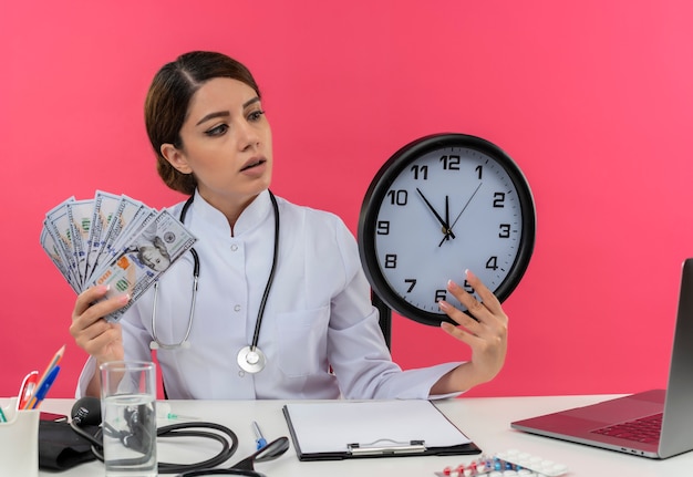 impressed young female doctor wearing medical robe and stethoscope sitting at desk with medical tools and laptop holding money and clock looking at clock 