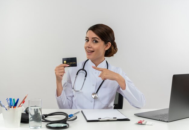 Impressed young female doctor wearing medical robe and stethoscope sitting at desk with medical tools and laptop holding and pointing at credit card isolated