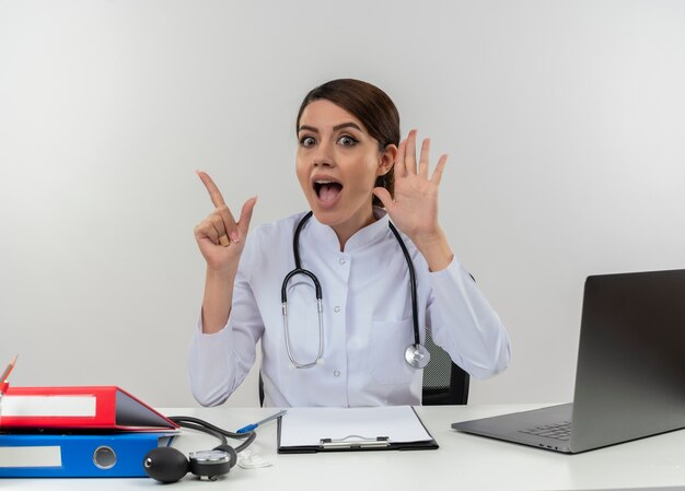 Impressed young female doctor wearing medical robe and stethoscope sitting at desk with medical tools and laptop keeping hand near ear raising finger isolated on white wall