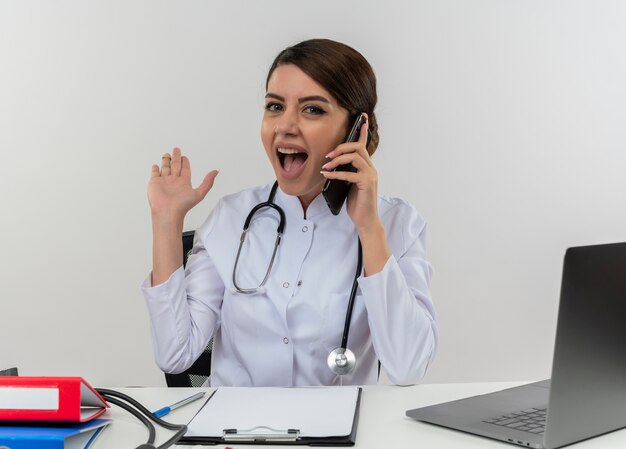 Impressed young female doctor wearing medical robe and stethoscope sitting at desk with medical tools and laptop talking on phone showing empty hand isolated on white wall