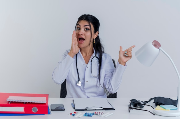 Impressed young female doctor wearing medical robe and stethoscope sitting at desk with medical tools looking pointing at side and whispering isolated