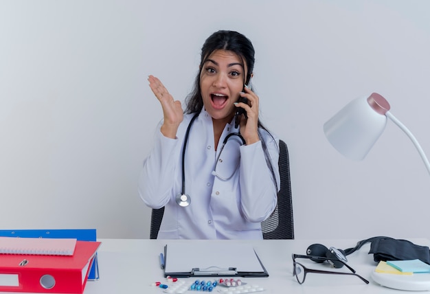 Impressed young female doctor wearing medical robe and stethoscope sitting at desk with medical tools looking talking on phone showing empty hand isolated