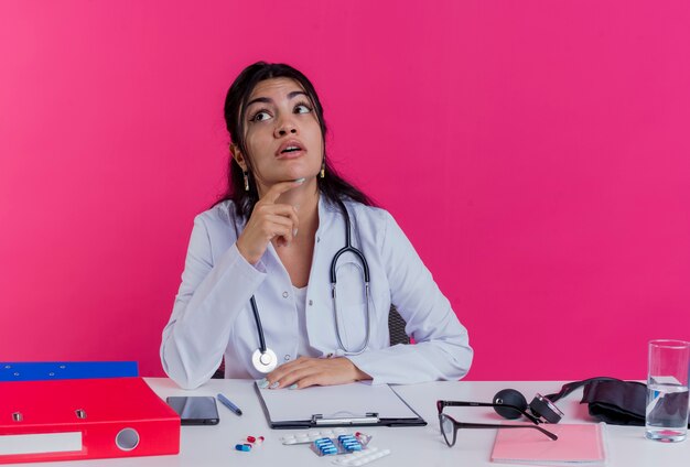 Впечатленная молодая женщина-врач в медицинском халате и стетоскопе, сидящая за столом с медицинскими инструментами, положив руку на стол, глядя в сторону и касаясь подбородка, изолированного на розовой стене