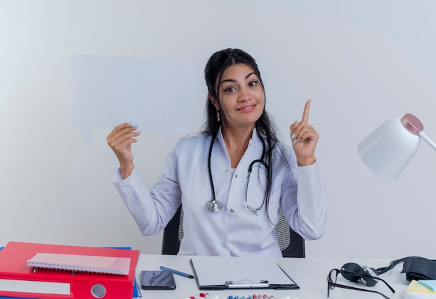 Foto gratuita giovane medico femminile impressionato che porta veste e stetoscopio medici che si siedono allo scrittorio con gli strumenti medici che alzano la bolla di chiacchierata della tenuta del dito che sembra isolata