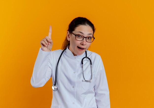 Impressed young female doctor wearing medical robe and stethoscope with glasses points at up isolated