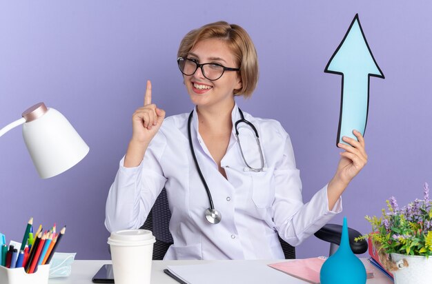 Impressionato giovane dottoressa che indossa abito medico con stetoscopio e occhiali si siede al tavolo con strumenti medici che tengono i punti del segno di direzione in alto isolato sulla parete blu