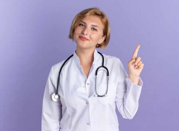 Впечатленная молодая женщина-врач в медицинском халате со стетоскопом указывает вверх, изолированную на синей стене