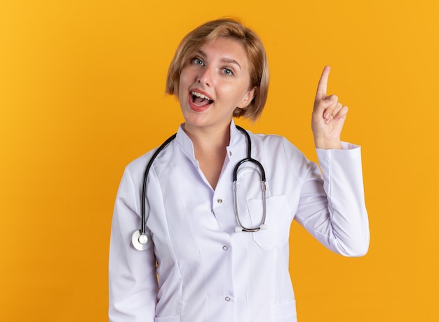オレンジ色の壁に隔離された聴診器のポイントで医療ローブを着ている感動の若い女性医師