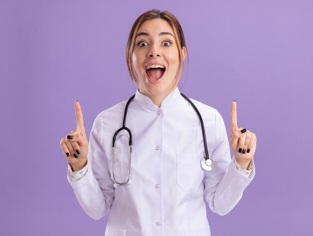 Впечатленная молодая женщина-врач в медицинском халате со стетоскопом указывает вверх, изолированную на фиолетовой стене с копией пространства