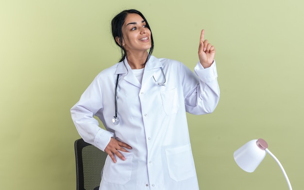 청진기가 올리브 녹색 벽에 고립 된 엉덩이 포인트에 손을 넣어 의료 가운을 입고 감동 된 젊은 여성 의사
