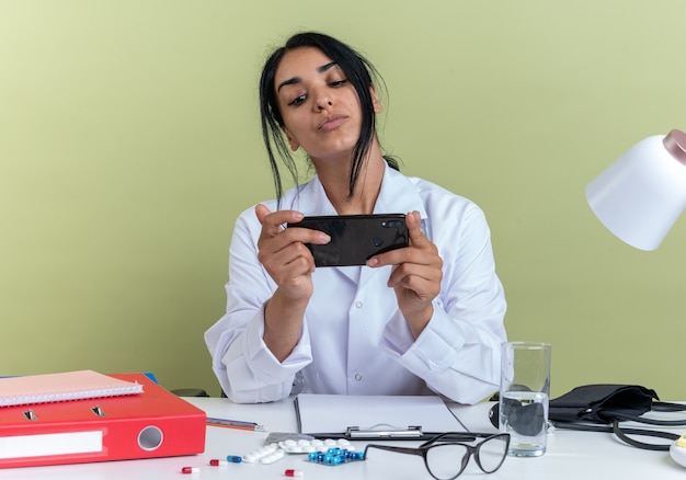 Una giovane dottoressa impressionata che indossa una veste medica con uno stetoscopio si siede alla scrivania con strumenti medici che gioca al telefono isolato su una parete verde oliva