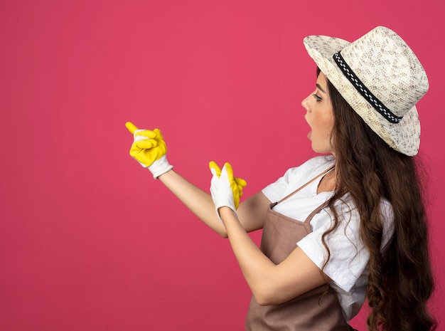 Impressed young female gardener in uniform wearing gardening hat and gloves looks and points at side isolated on pink wall with copy space