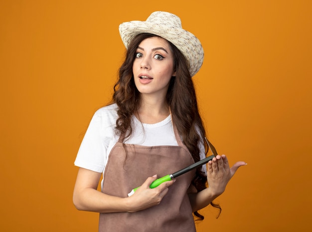 Impressed young female gardener in uniform wearing gardening hat holds hoe rake isolated on orange wall with copy space