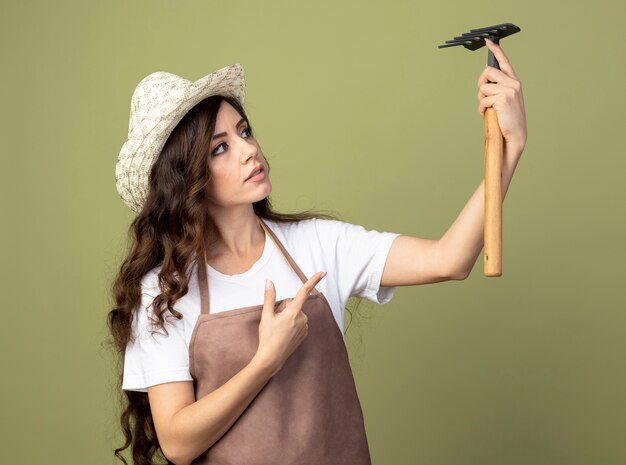 Impressed young female gardener in uniform wearing gardening hat holds and points at rake isolated on olive green wall