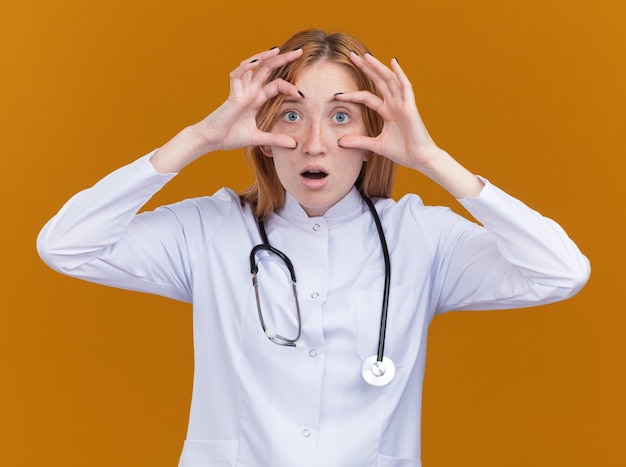 Impressed young female ginger doctor wearing medical robe and stethoscope doing look gesture using hands as binoculars