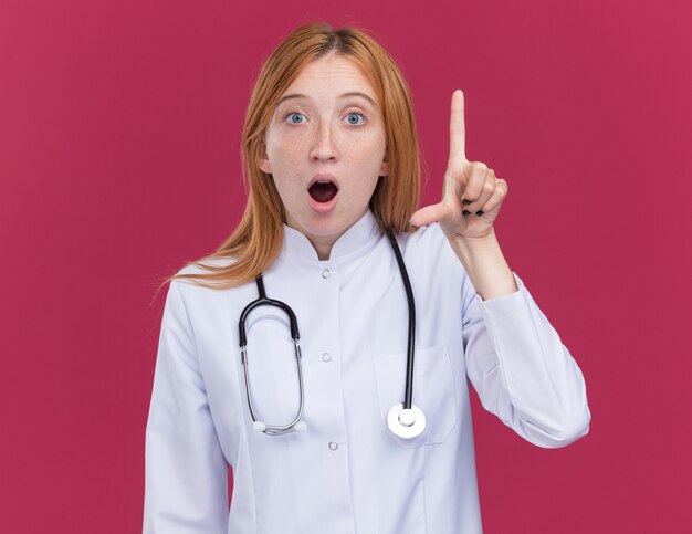 敗者のジェスチャーをしている医療ローブと聴診器を身に着けている感銘を受けた若い女性の生姜医師