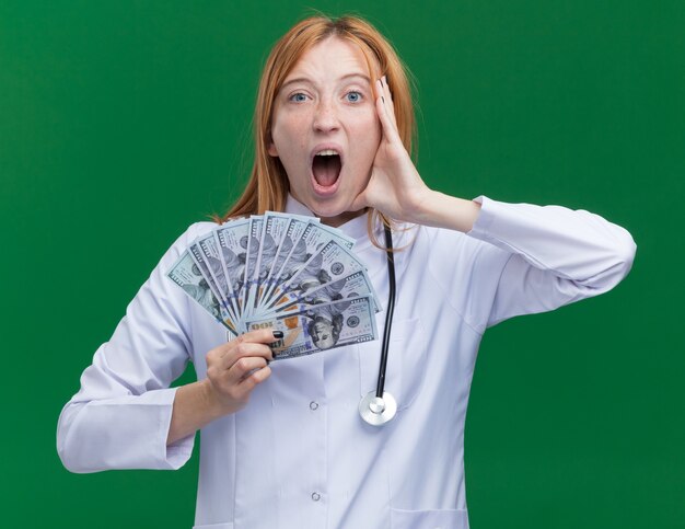 Impressed young female ginger doctor wearing medical robe and stethoscope holding money touching face looking at front screaming isolated on green wall