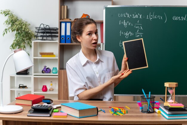 教室の前を見てミニ黒板を持って指さしている学用品を持って机に座っている感銘を受けた若い女性の数学の先生