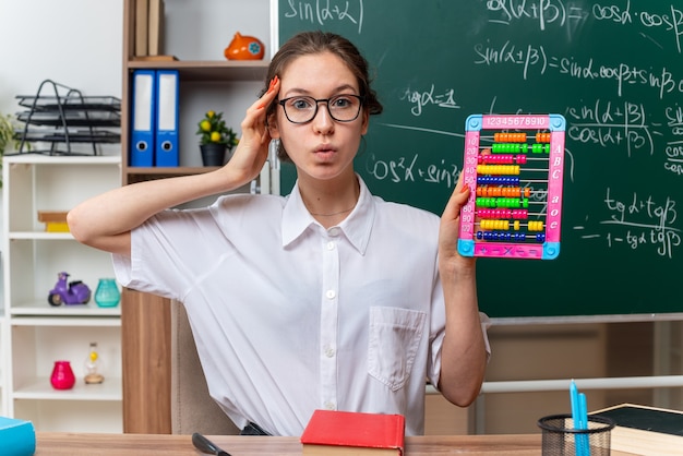 Foto gratuita impressionato giovane insegnante di matematica femminile con gli occhiali seduto alla scrivania con materiale scolastico che mostra l'abaco guardando davanti tenendo la mano sulla testa in classe