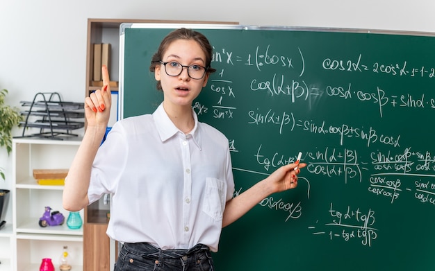 impressed young female math teacher wearing glasses standing in front of chalkboard looking at front pointing with chalk at math problem written on chalkboard pointing up in classroom