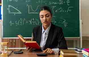 Free photo impressed young female teacher sits at table with school supplies holding book and looking at calculator in her hand in classroom