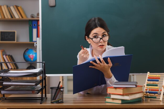 教室で学校の道具を使って机に座って鉛筆で本を持って眼鏡をかけている感動の若い女性教師