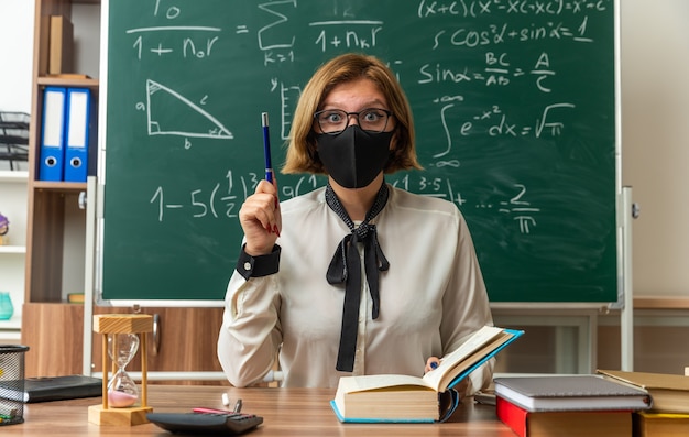 Impressed young female teacher wearing glasses and medical mask sits at table with school tools holding pen in classroom