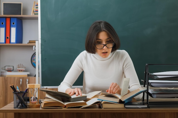 Impressionato giovane insegnante femminile con gli occhiali che legge un libro seduto alla scrivania con gli strumenti della scuola in classe