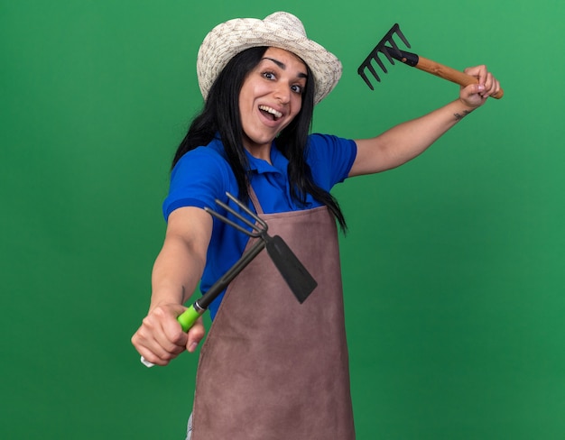 Free photo impressed young gardener girl wearing uniform and hat raising rake and stretching out hoe-rake towards front looking at front isolated on green wall