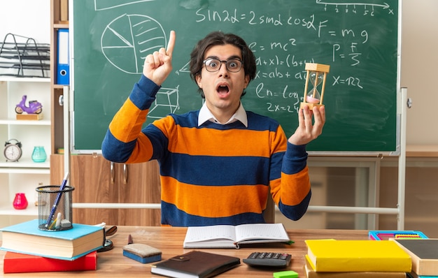 impressed young geometry teacher wearing glasses sitting at desk with school supplies in classroom holding hourglass looking at front pointing up