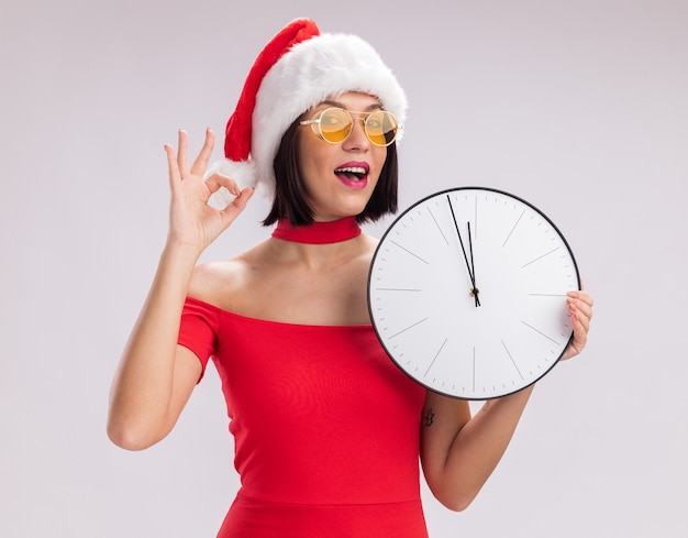 산타 모자와 흰색 배경에 고립 된 확인 표시를 하 고 카메라를보고 시계를 들고 안경을 착용하는 감동 어린 소녀