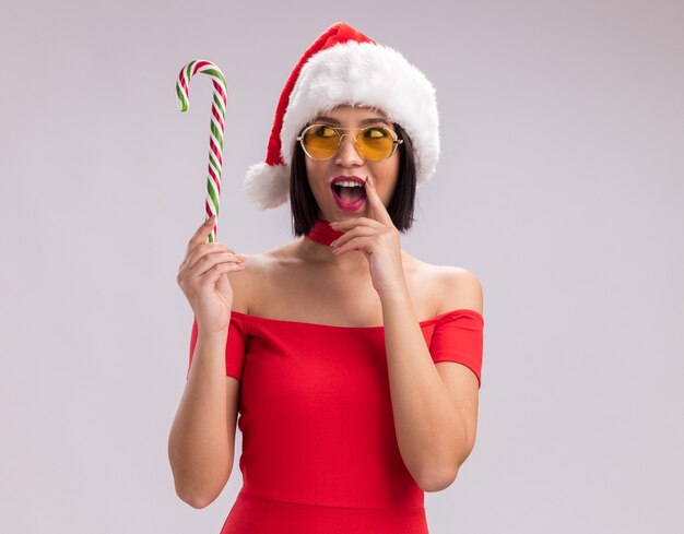 Impressed young girl wearing santa hat and glasses holding and looking at christmas candy cane touching lip isolated on white background with copy space