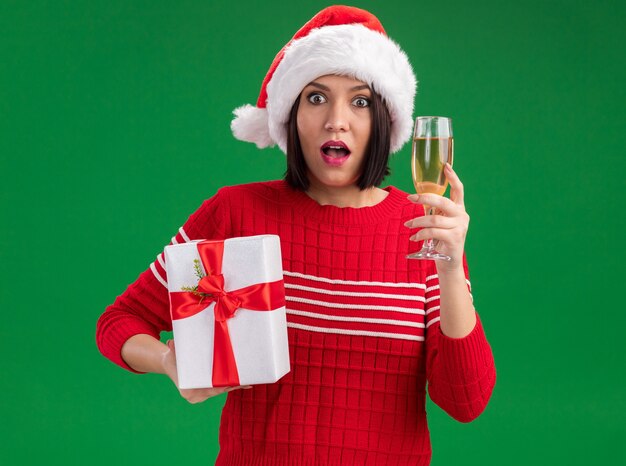 Impressed young girl wearing santa hat holding gift package and glass of champagne  isolated on green wall