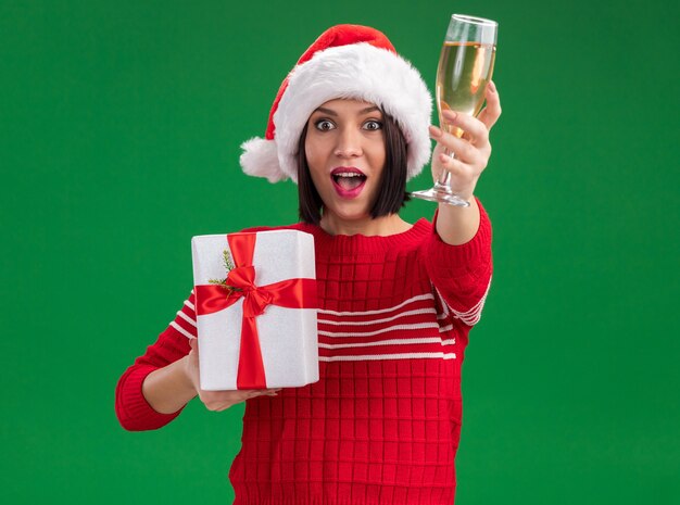 Впечатленная молодая девушка в шляпе санта-клауса держит подарочный пакет и протягивает бокал шампанского, изолированного на зеленой стене