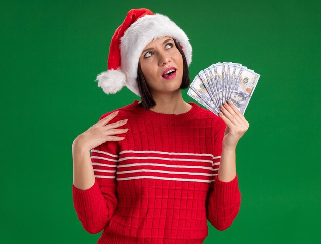 Impressed young girl wearing santa hat holding money touching shoulder looking up isolated on green wall