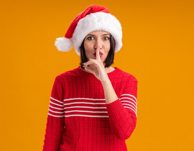 Impressed young girl wearing santa hat looking at camera doing silence gesture isolated on orange background with copy space