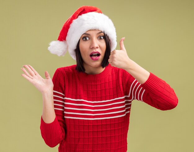 Impressed young girl wearing santa hat looking at camera showing empty hand and thumb up isolated on olive green background