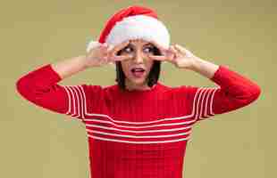 Free photo impressed young girl wearing santa hat looking at side showing v-sign symbols near eyes isolated on olive green wall