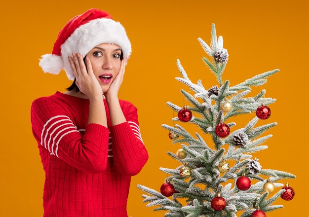 Впечатленная молодая девушка в шляпе санта-клауса, стоящая возле украшенной рождественской елки, держа руки на лице, глядя в камеру, изолированную на оранжевом фоне