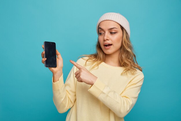 Impressed young girl wearing winter hat holding looking and pointing at mobile phone 