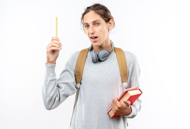 Impressed young guy student wearing backpack with headphones on neck holding books raising pen isolated on white wall