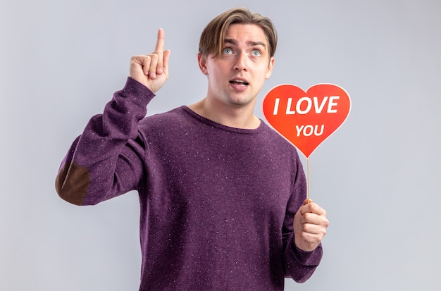 Free photo impressed young guy on valentines day holding red heart on a stick with i love you text points at up isolated on white background