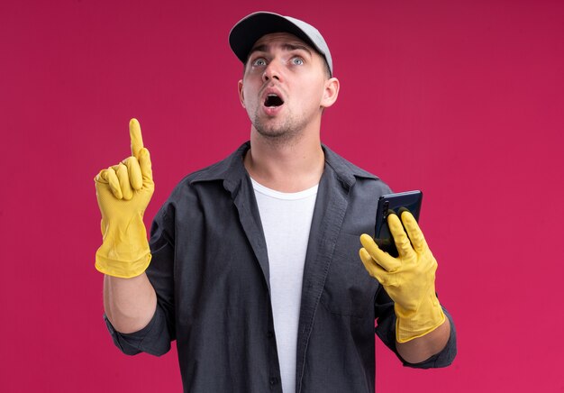 ピンクの壁に隔離された上で電話ポイントを保持している手袋でTシャツとキャップを身に着けている感動の若い厄介な掃除人