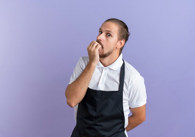 Impressed young handsome barber wearing uniform biting his fingers and keeping hand behind back isolated on purple  with copy space